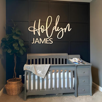 Nursery room with a gray crib, dark wall with 'Holdyn James' sign, and a plant.