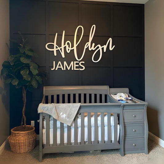 Nursery room with a gray crib, dark wall with 'Holdyn James' sign, and a plant.